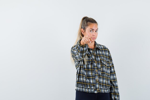 Señorita apuntando a la cámara en camisa, pantalones cortos y mirando confiada, vista frontal.