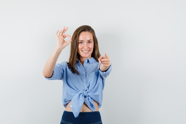 Señorita apuntando a la cámara con signo ok en camisa azul, pantalones y mirando contento