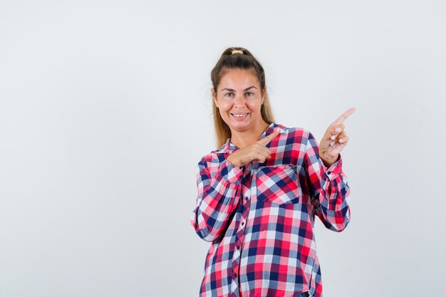 Señorita apuntando a la esquina superior derecha con camisa a cuadros y mirando alegre, vista frontal.