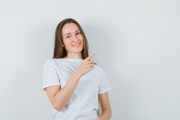 Señorita apuntando a la esquina superior derecha en camiseta blanca y mirando alegre