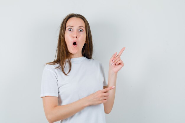 Señorita apuntando a la esquina superior derecha en camiseta blanca y mirando sorprendido