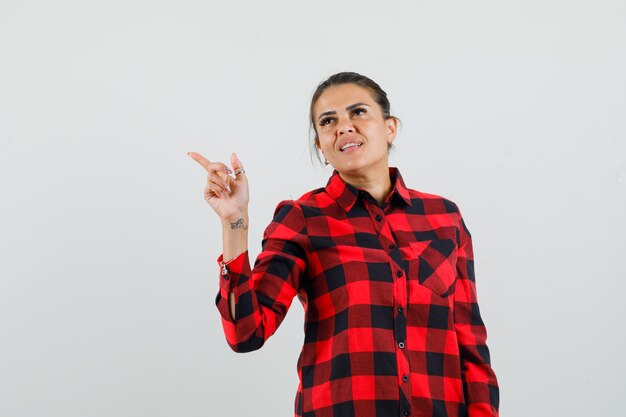 Señorita apuntando a la esquina superior izquierda en camisa a cuadros y mirando esperanzada.