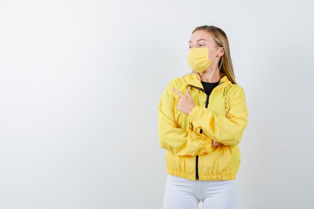 Señorita apuntando a la esquina superior izquierda en chaqueta, pantalón, máscara y luciendo confiada. vista frontal.