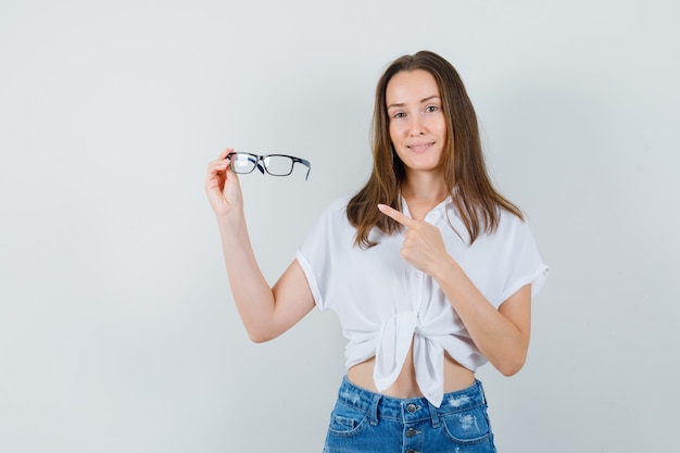 Señorita apuntando a gafas en blusa blanca y mirando positivo. vista frontal.