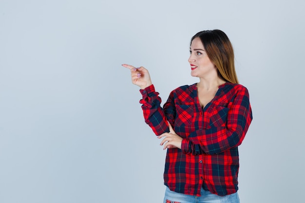 Señorita apuntando a un lado con camisa a cuadros y mirando asombrado, vista frontal.