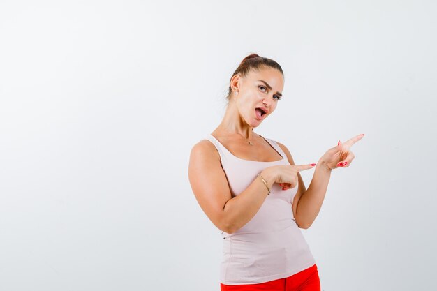 Señorita apuntando a un lado en la camiseta sin mangas beige y luciendo atractiva. vista frontal.