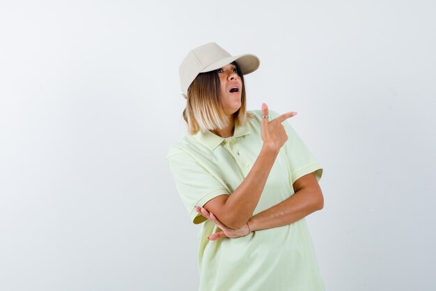 Señorita apuntando hacia el lado derecho en camiseta, gorra y mirando perpleja. vista frontal.