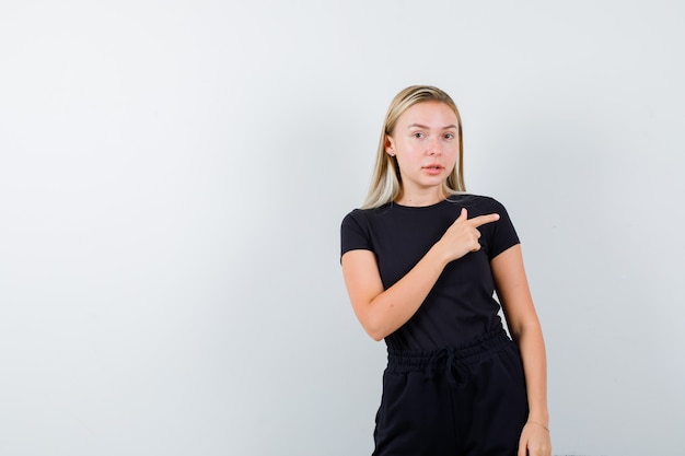 Señorita apuntando hacia el lado derecho en camiseta, pantalón y mirando enfocado. vista frontal.