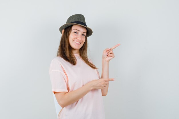 Señorita apuntando hacia el lado derecho en camiseta rosa, sombrero y mirando alegre. vista frontal.