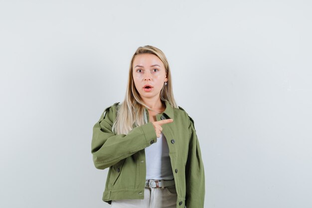 Señorita apuntando hacia el lado derecho en chaqueta, camiseta y mirando sorprendido. vista frontal.