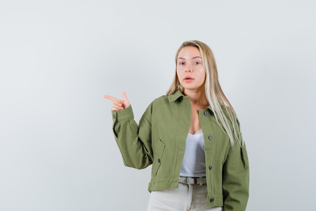 Señorita apuntando hacia el lado derecho en chaqueta, pantalones y mirando vacilante. vista frontal.