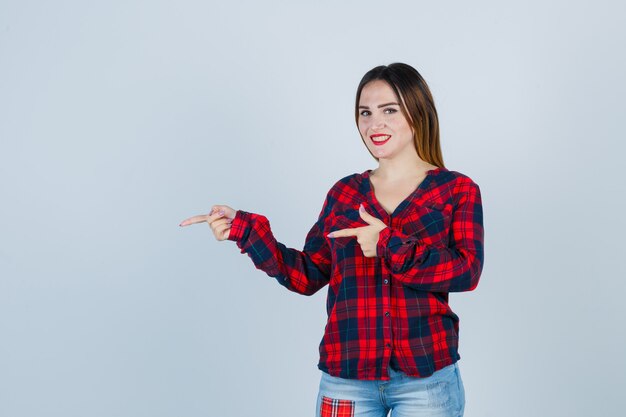 Señorita apuntando hacia el lado izquierdo en camisa a cuadros, jeans y mirando alegre, vista frontal.