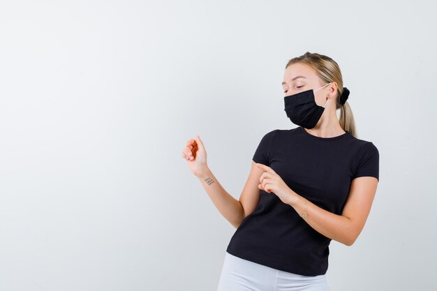 Señorita apuntando hacia el lado izquierdo en camiseta, pantalón, máscara médica y mirando desconcertado