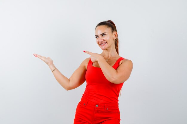 Señorita apuntando hacia el lado izquierdo en camiseta roja, pantalón rojo y mirando alegre, vista frontal.