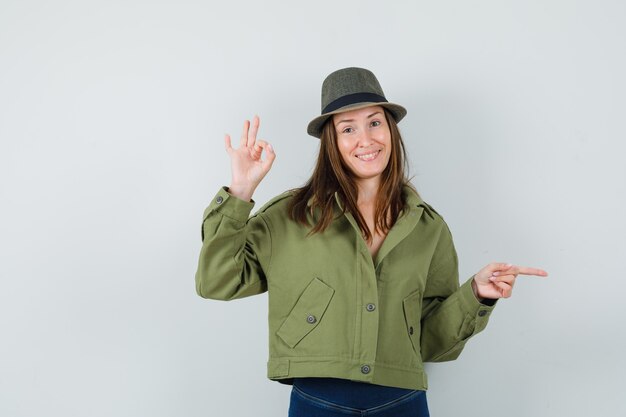 Señorita apuntando a un lado mostrando ok firmar en chaqueta pantalones sombrero y mirando alegre