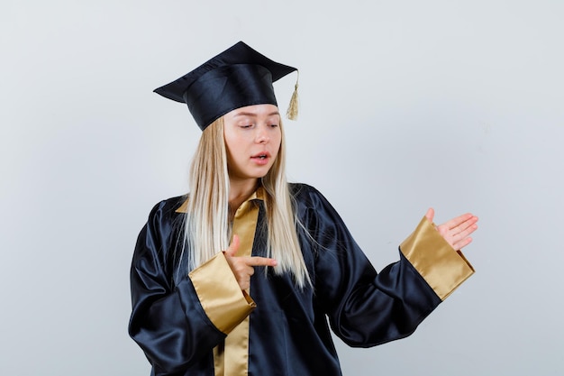 Señorita apuntando a un lado en traje académico y mirando confiada.