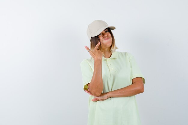 Señorita apuntando a las mejillas sopladas en camiseta, gorra y con aspecto divertido. vista frontal.