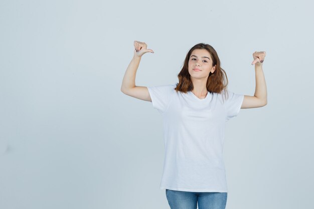 Señorita apuntando a sí misma con los pulgares en camiseta, jeans y luciendo orgullosa, vista frontal.