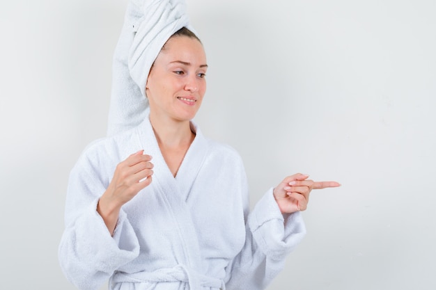 Señorita en bata de baño blanca, toalla apuntando hacia el lado derecho y mirando alegre, vista frontal.