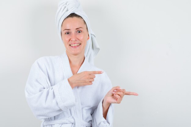 Señorita en bata de baño blanca, toalla apuntando a un lado y mirando alegre, vista frontal.