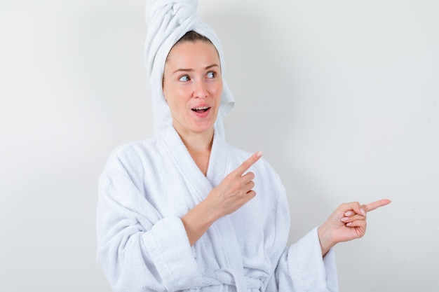 Señorita en bata de baño blanca, toalla apuntando a un lado y mirando asombrado, vista frontal.