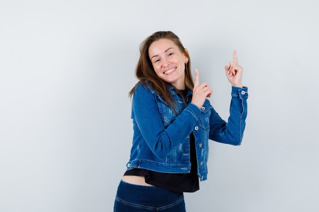 Señorita en blusa, chaqueta apuntando hacia arriba y mirando alegre, vista frontal.