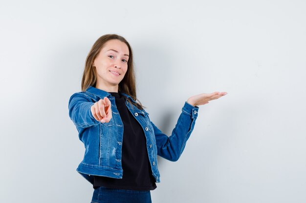 Señorita en blusa, chaqueta apuntando a la cámara, mostrando algo y mirando confiado, vista frontal.