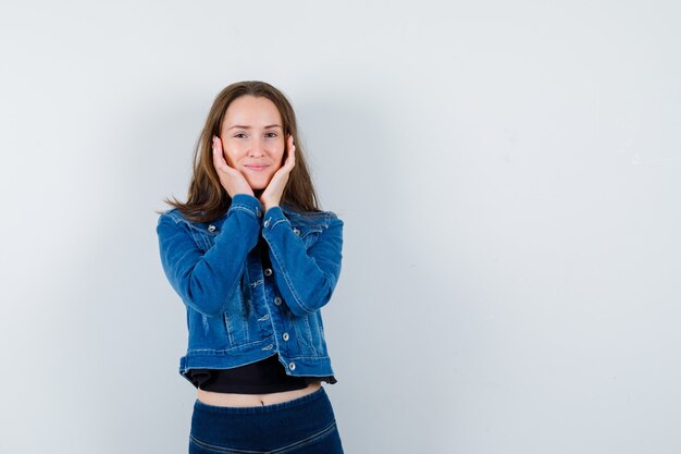 Señorita en blusa, chaqueta cogidos de la mano en las mejillas y luciendo elegante, vista frontal.