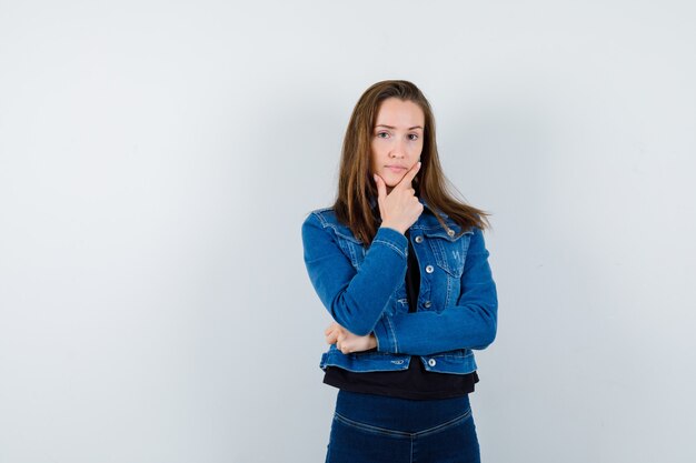 Señorita en blusa, chaqueta, jeans sosteniendo su barbilla mientras piensa y se ve sensible, vista frontal.