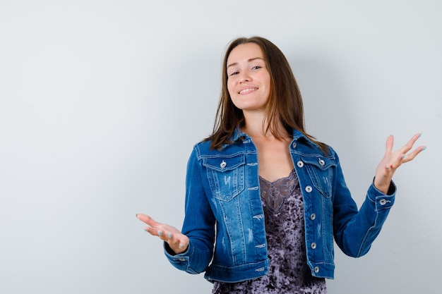 Señorita en blusa, chaqueta de mezclilla pretendiendo sostener algo y mirando feliz, vista frontal.