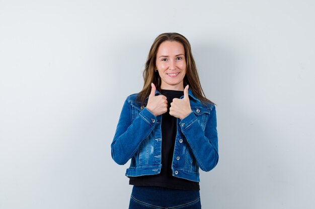 Señorita en blusa, chaqueta mostrando doble pulgar hacia arriba y mirando confiado, vista frontal.