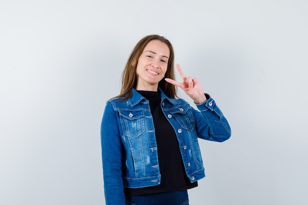 Señorita en blusa, chaqueta mostrando el signo V y mirando alegre, vista frontal.