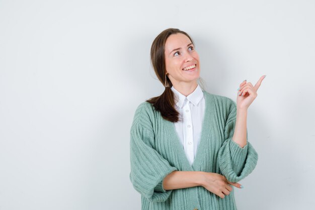 Señorita en blusa, chaqueta de punto apuntando a la esquina superior derecha, mirando hacia arriba y mirando jovial, vista frontal.