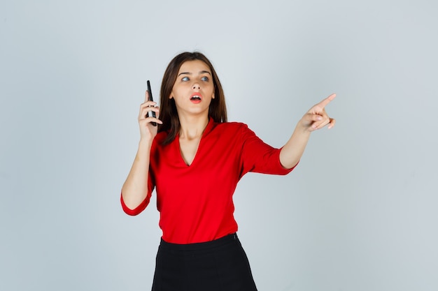 Señorita en blusa roja, falda hablando por teléfono móvil mientras apunta a un lado