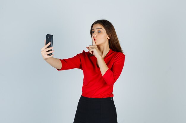 Señorita en blusa roja, falda haciendo videollamadas mientras muestra gesto de silencio