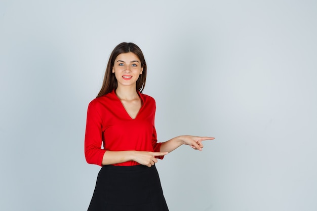 Señorita en blusa roja, falda negra apuntando hacia la derecha con los dedos índices y mirando alegre