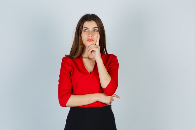 Señorita en blusa roja, falda de pie en pose de pensamiento y mirando pensativo