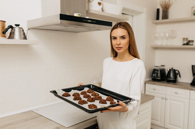 Señorita con cabello castaño claro en camisa blanca cocinar galletas de chocolate caseras en cocina ligera