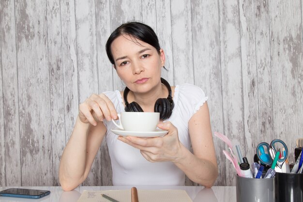 Señorita en camisa blanca tomando café escuchando música en auriculares negros escribiendo notas en la pared gris