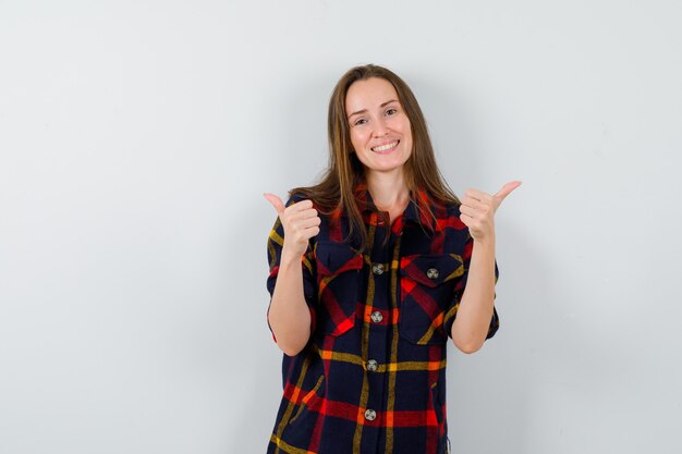 Señorita en camisa casual mostrando doble pulgar hacia arriba y mirando alegre, vista frontal.