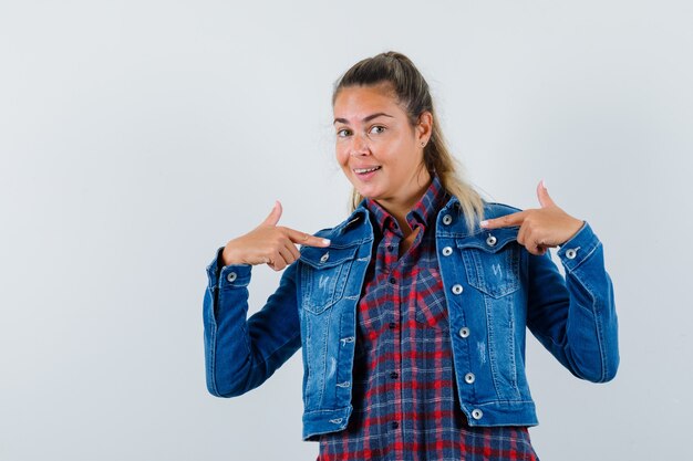 Señorita en camisa, chaqueta apuntando a sí misma y mirando alegre, vista frontal.