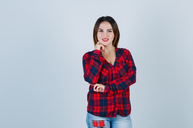 Señorita en camisa a cuadros apoyando la barbilla en la mano y mirando alegre, vista frontal.