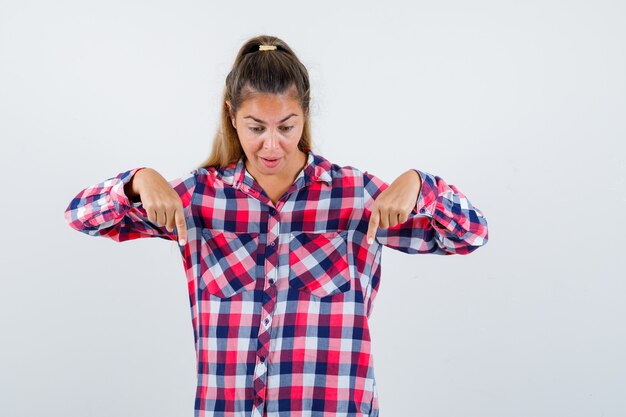 Señorita en camisa a cuadros apuntando hacia abajo y mirando asombrado, vista frontal.