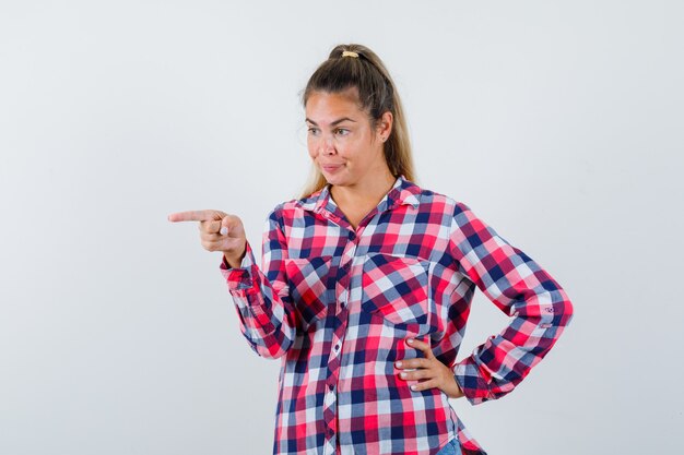 Señorita en camisa a cuadros apuntando a algo lejos y mirando alegre, vista frontal.