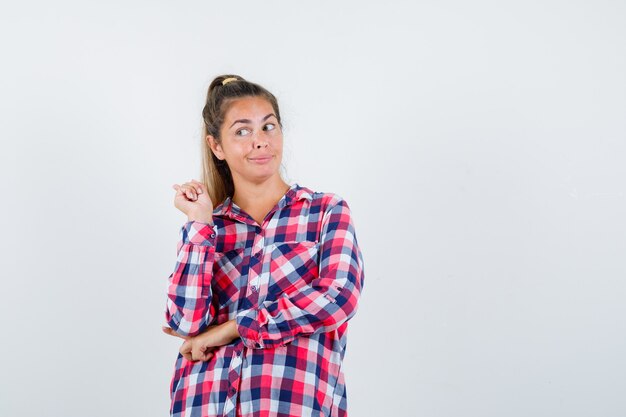 Señorita en camisa a cuadros apuntando hacia atrás y mirando curiosa, vista frontal.