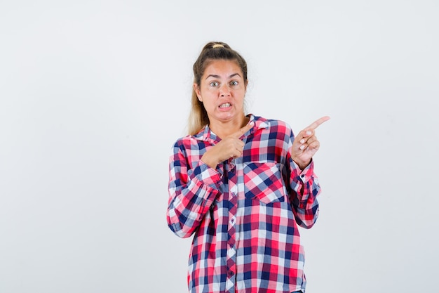 Señorita en camisa a cuadros apuntando hacia el lado derecho y mirando preocupada, vista frontal.