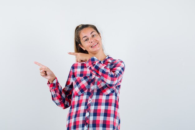 Señorita en camisa a cuadros apuntando hacia el lado izquierdo y mirando feliz, vista frontal.