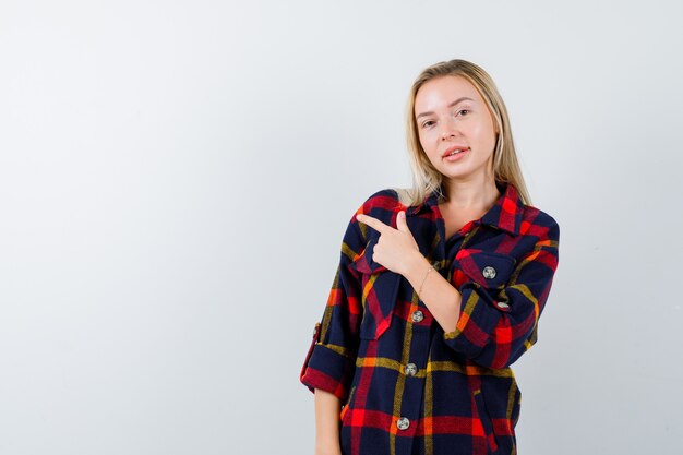 Señorita en camisa a cuadros apuntando hacia el lado izquierdo y mirando feliz, vista frontal.