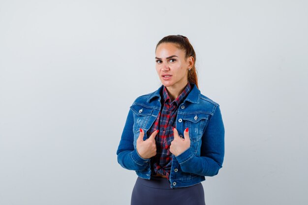 Señorita en camisa a cuadros, chaqueta de mezclilla apuntando a sí misma y mirando con cuidado, vista frontal.