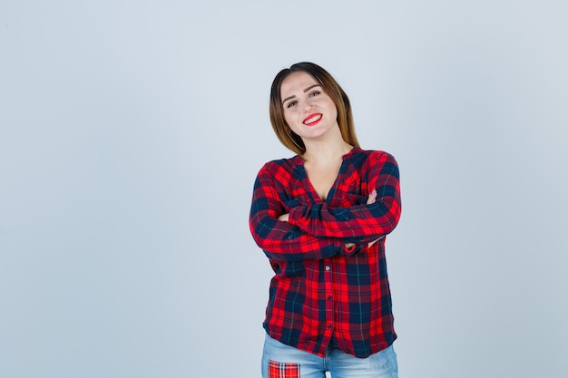 Señorita en camisa a cuadros sosteniendo los brazos cruzados y mirando alegre, vista frontal.